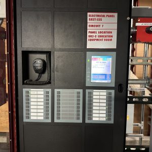 Gamewell FCI fire alarm control panel with voice-evacuation mic, LCD status screen, and annunciators installed in a hospital equipment room.