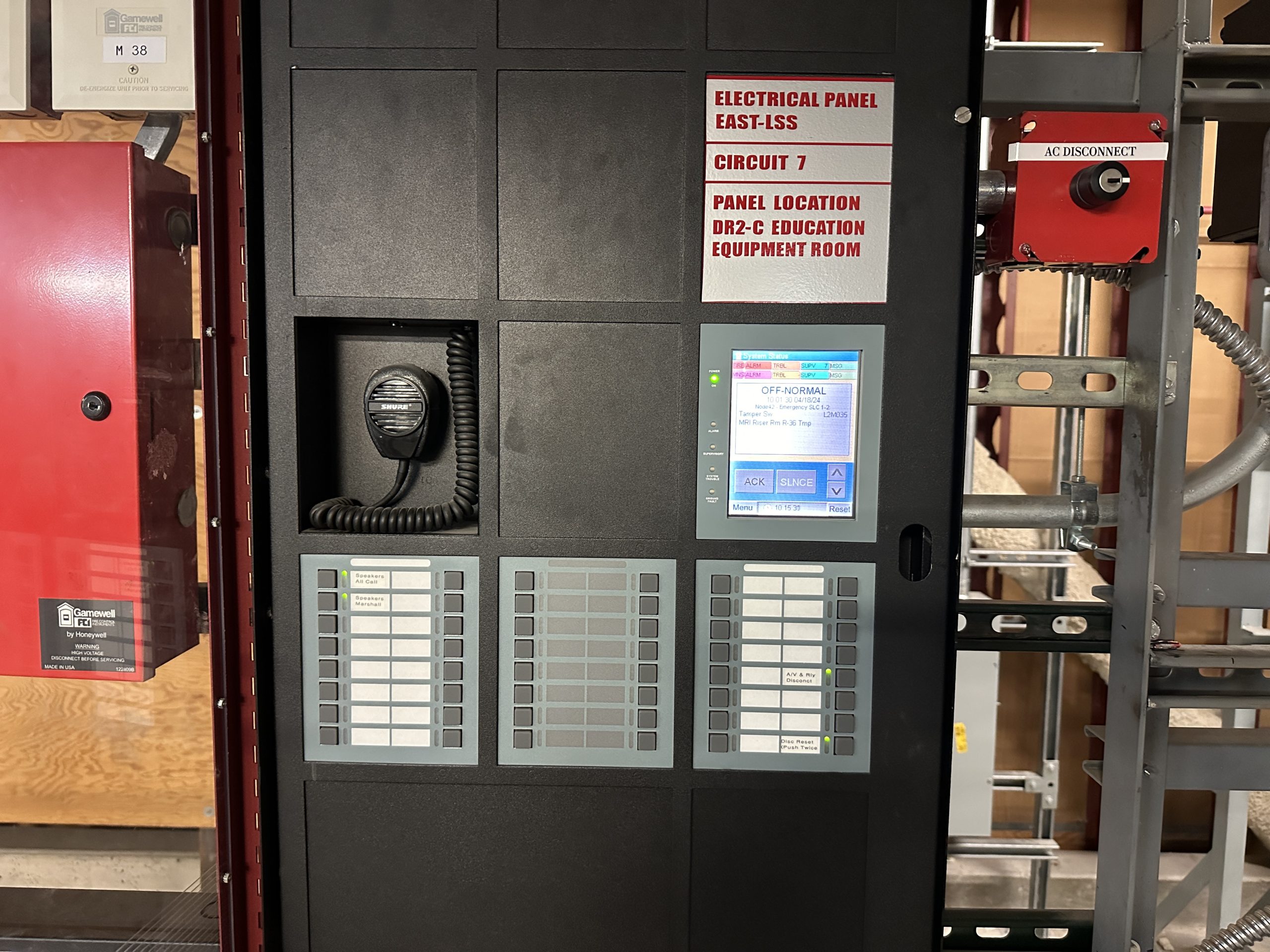 Gamewell FCI fire alarm control panel with voice-evacuation mic, LCD status screen, and annunciators installed in a hospital equipment room.